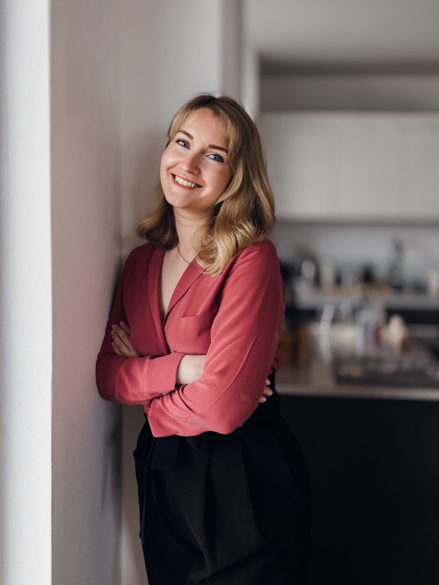 Anna Buldakova smiling, arms crossed, leaning against a wall at home